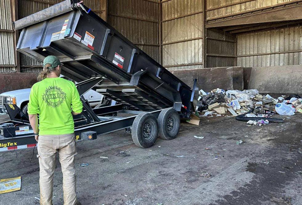 Image of a man in a green shirt that reads "Garity's Grass" standing in front of a trailer dumping trash into a large dump area.
